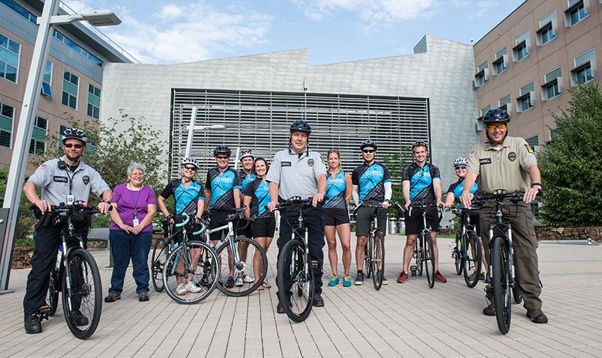 A group of cyclists, some with blue jerseys and some with security uniforms, stand with their bikes in front of a modern building