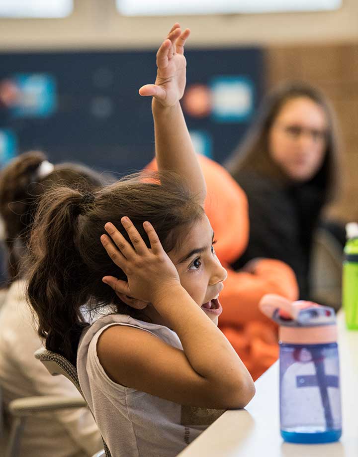 A young girl eagerly raises her hand in a classroom.