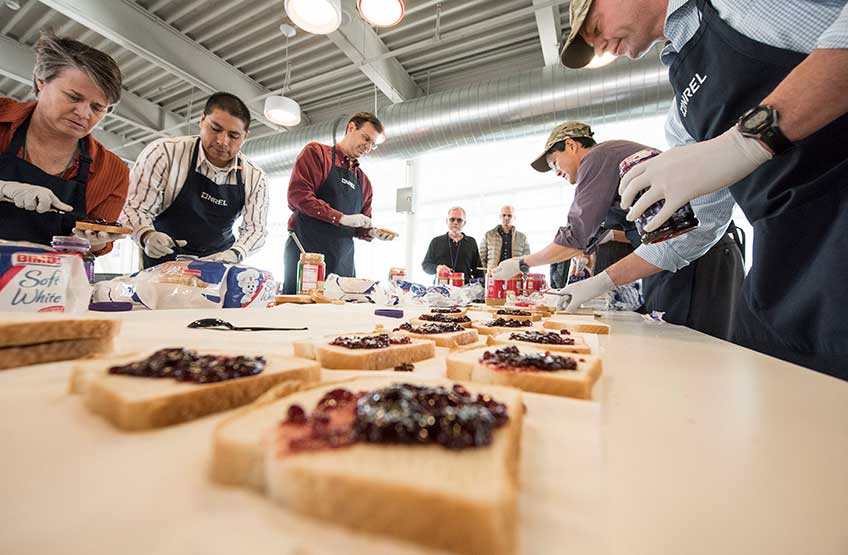 A group of people standing on opposite sides of a table make peanut butter and jelly sandwiches.