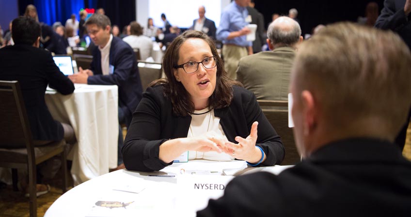 Woman sitting at a table in a conference area having a discussion with a person facing away from the camera