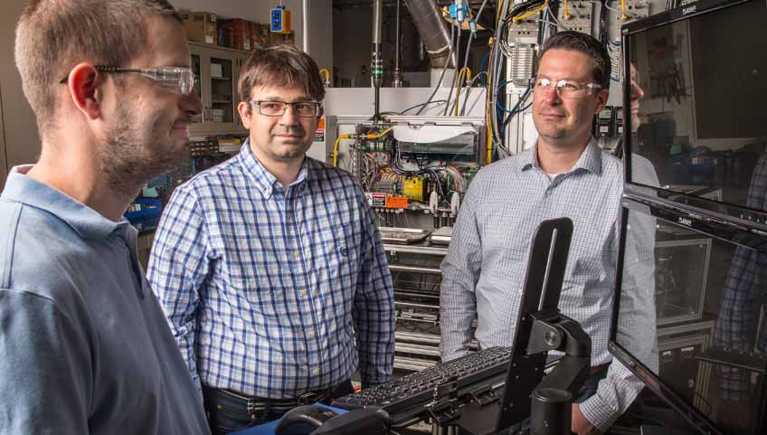 Three men stand next to a piece of laboratory equipment.