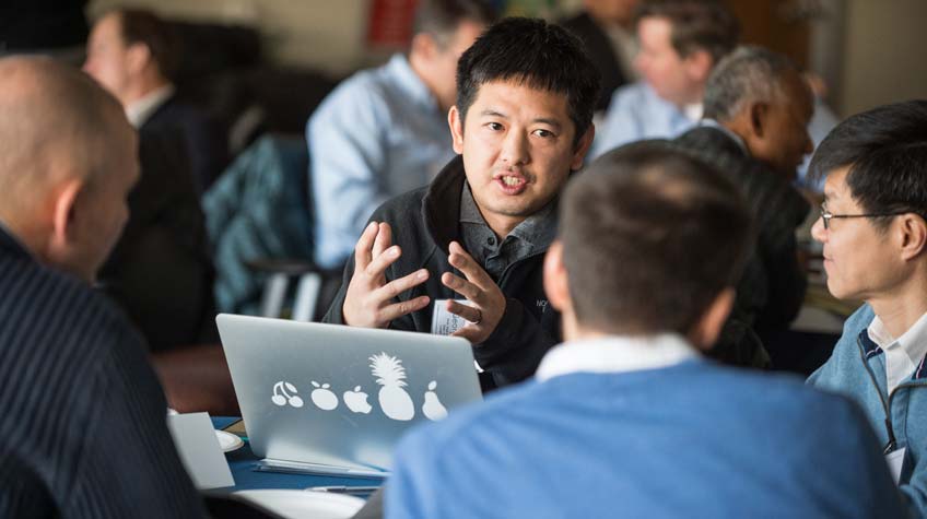 Photo of four men sitting at a table and having a discussion. One of the men has an open laptop in front of him.