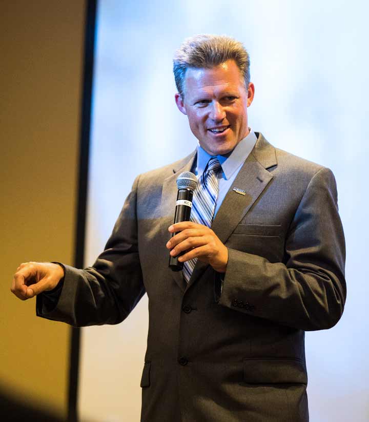 Photo of a man speaking into a microphone in front of a screen.