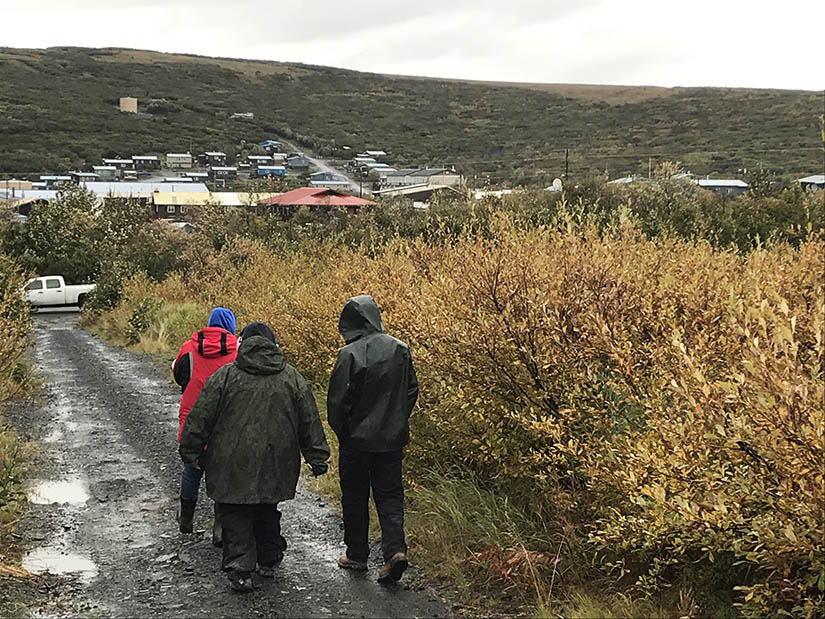 Three people walking on a muddy road to Mountain Village, Alaska, on a hill in the background