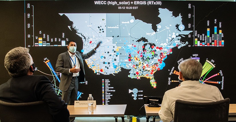 A researcher standing at a large visualization screen speaks to two seated people viewing.