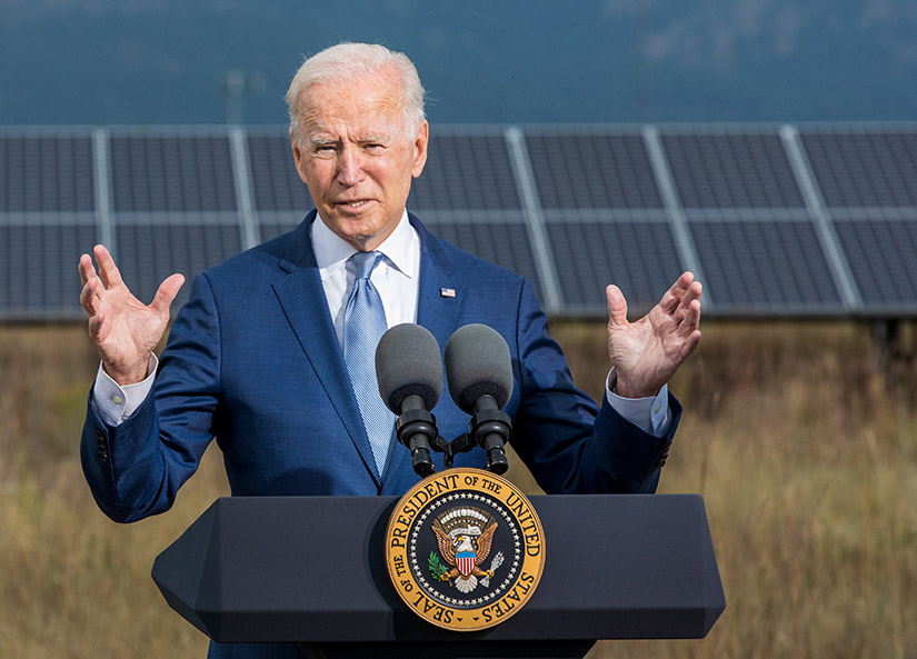 A person in a suit and tie standing at a podium with solar panels and mountains in the background