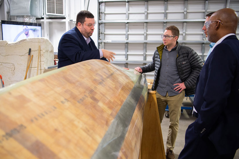 Photo of four people standing in a workshop, a large wind turbine blade is in the foreground.