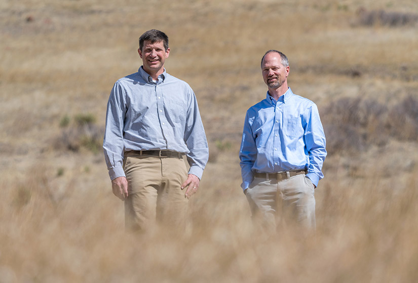 Photo of two men standing outside in a field of tall brown grass