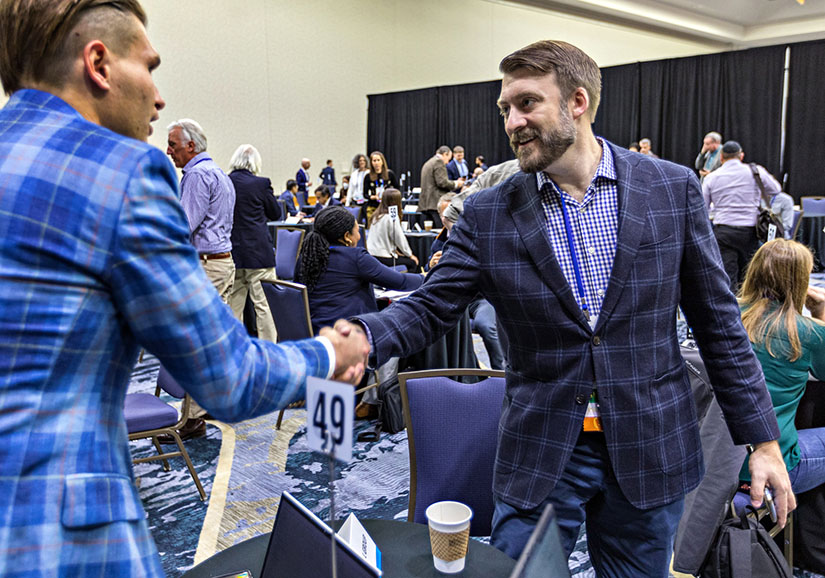 Two people shake hands in a large room full of people at meeting tables