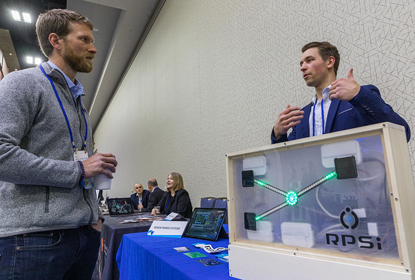 Two people speak at an exhibitor table at an event with an electrical device on the table