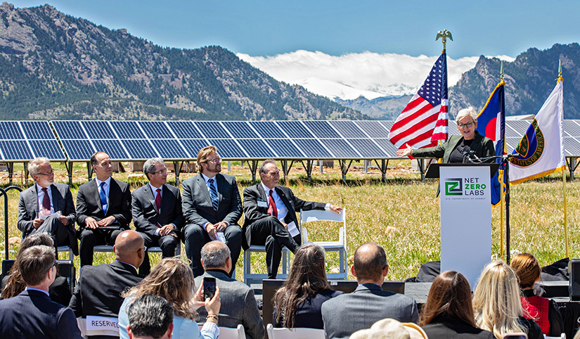 Photo of Secretary of Energy Jennifer Granholm speaking at NREL's Flatirons Campus with other speakers on stage and a seated audience with flags, mountains, and solar panels in the background