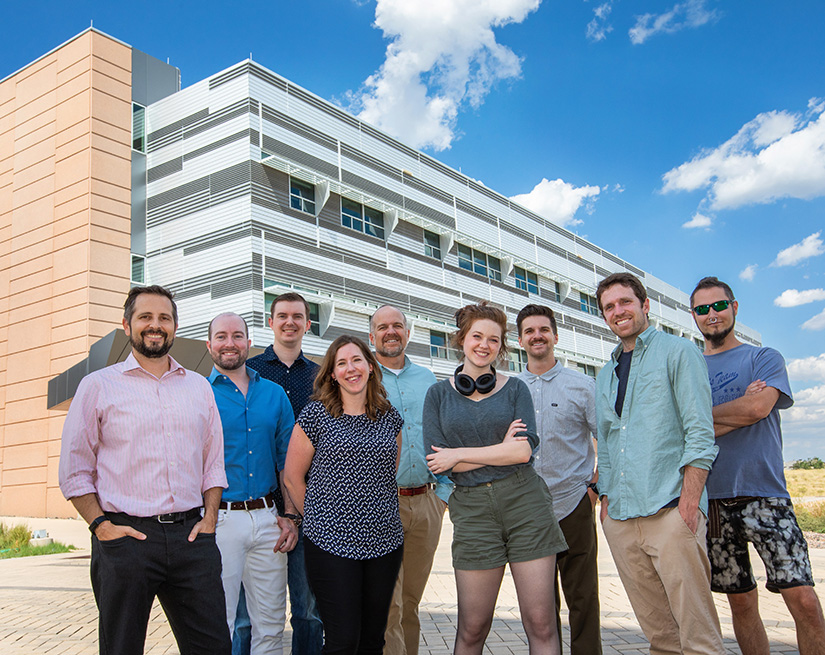 Photo of nine smiling people standing outside of a research facility building under a blue sky