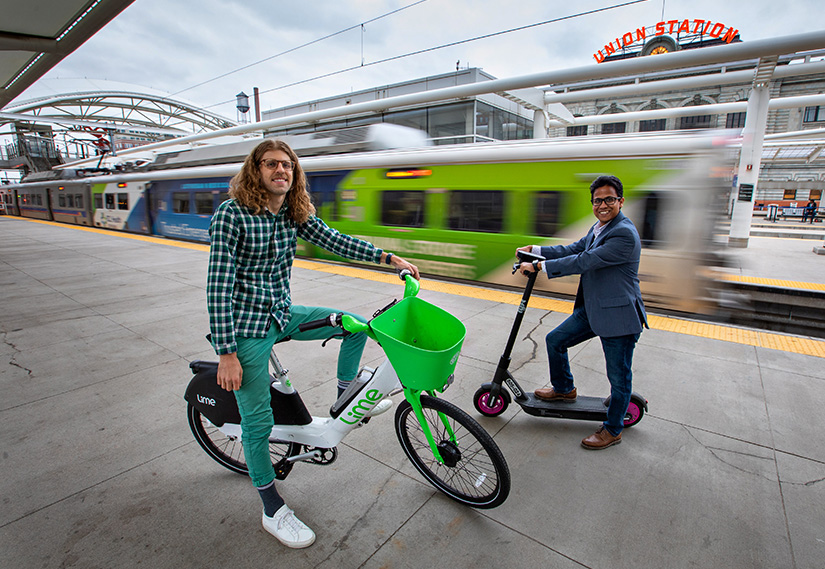 Dos personas, una en una bicicleta eléctrica de viaje compartido y la otra en un scooter eléctrico de viaje compartido, se paran en una plataforma de tren