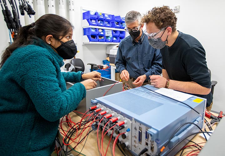 Three people work together in a laboratory connecting wires to metal boxes, preparing the equipment for testing.