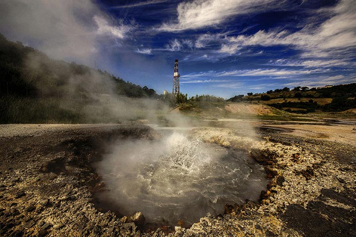 Photo of a steaming pool of water in an outdoor setting with drilling rig in the background.