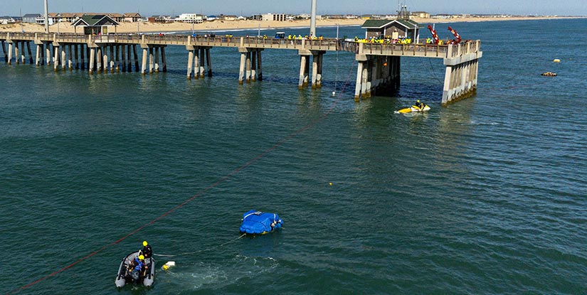 Overhead shot of a small boat in the ocean tethered to a floating piece of equipment, with a pier full of people in the background.