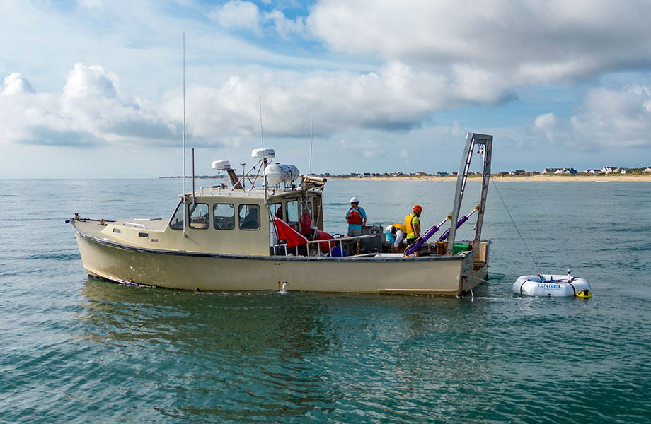 A boat and crew with the WEC device in the water behind it. The shoreline is seen in the distance.