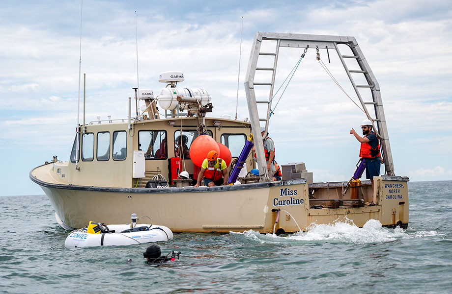 Divers in the sea with the WEC device behind the boat.