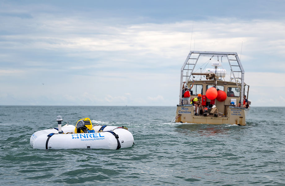 The WEC device floats in the foreground as the boat and crew float in the background.
