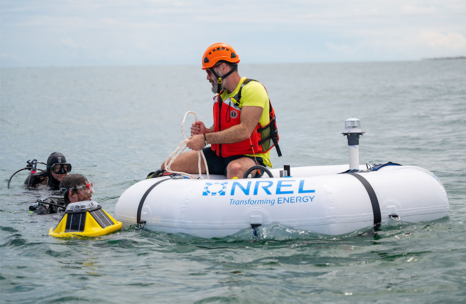 A man sits on the WEC device in the sea, talking to two divers in the water.