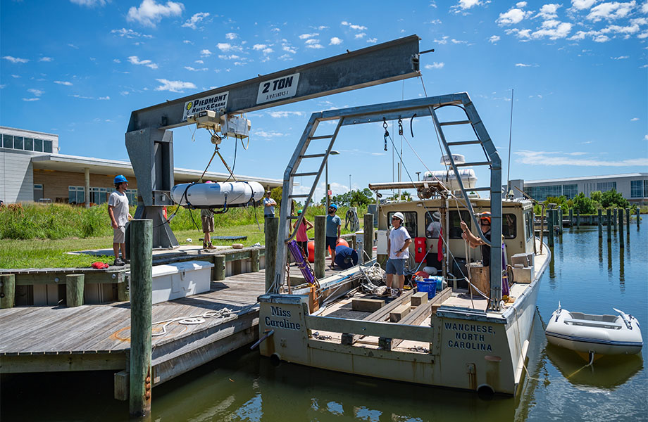 A crane hold the WEC device over a pier as crewe watch. A boat floats next to the pier.