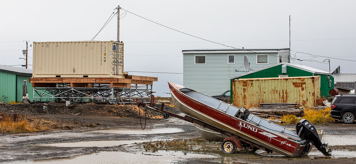 A new, elevated container home sits in front of older buildings and a boat.