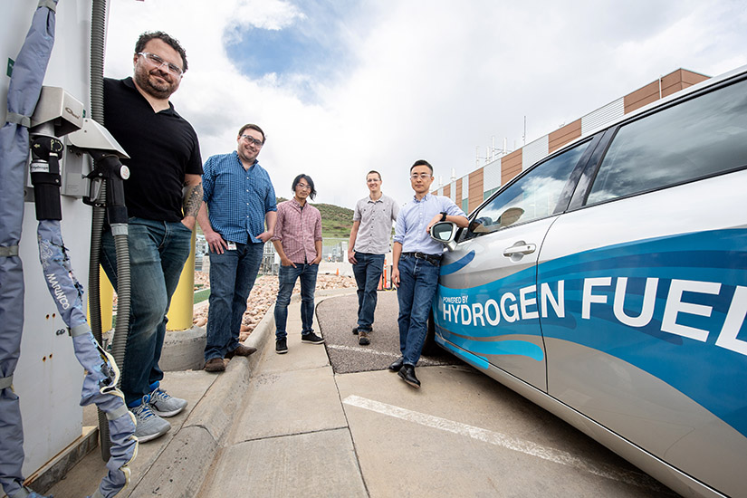 A group stands in between a fuel pump and a car that has hydrogen fuel written on it.