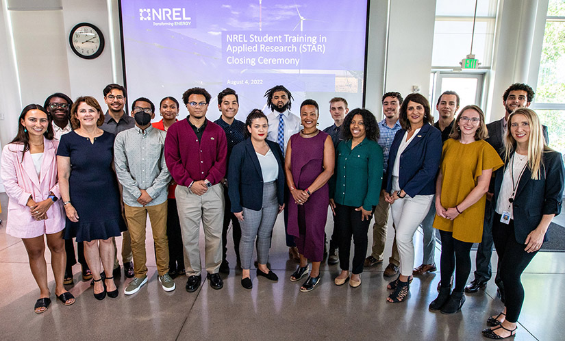19 people standing and posing for group photo at NREL in front of a projected slide image that reads: "NREL Student Training in Applied Research (STAR) Closing Ceremony. August 4, 2022"