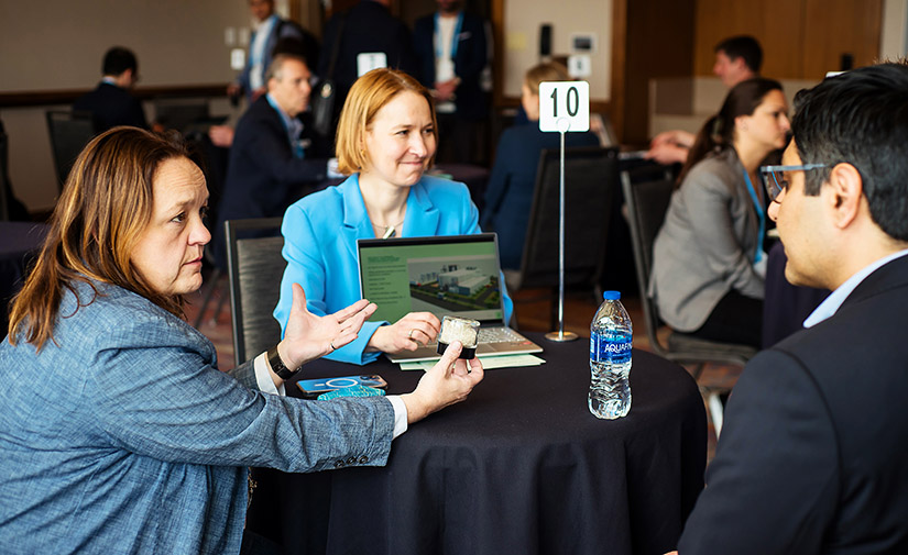 Two women sit at a round table with a man. One woman holds up something while the other shows off a laptop screen.