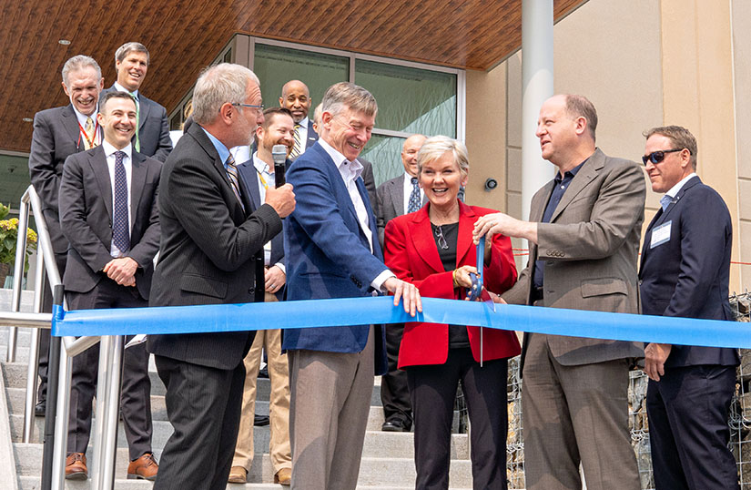 A group of people stand on the steps of a building to cut a ribbon with giant scissors.