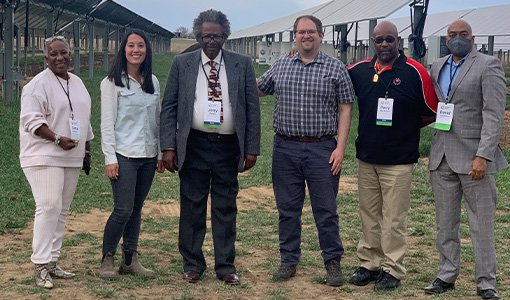 Six people stand in front of commercial-scale solar panels on a farm.