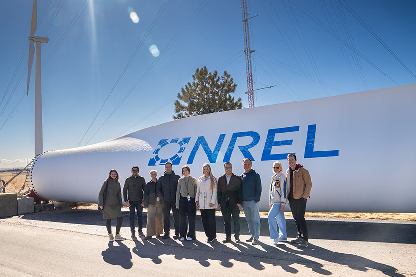 A group of people stand in front of a wind turbine with the letters NREL inscribed.