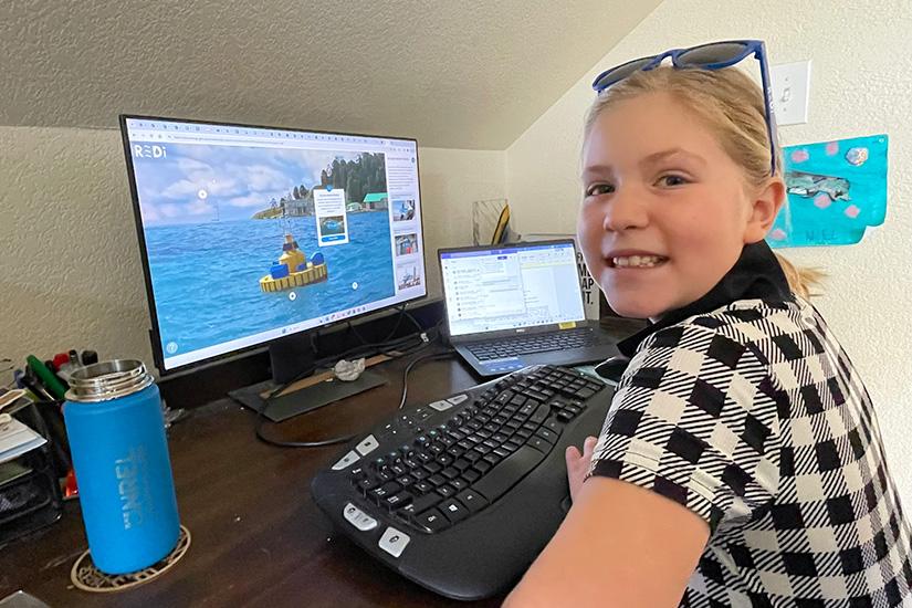 A young girl smiles at the camera as she sits at a computer with the REDI Island application on the screen