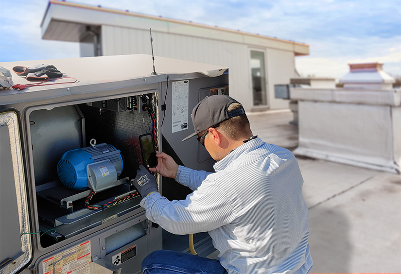 A man installs a blue motor in an HVAC unit on the roof of a building.