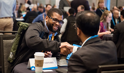 In a room full of people at tables, two men sitting at a table shake hands. 