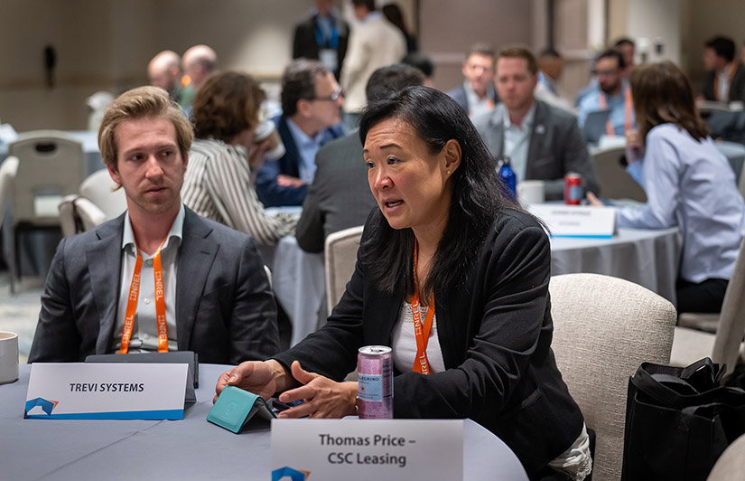 A woman sits at a round table next to a man speaking to someone off camera. Other people sit at additional round tables behind her.