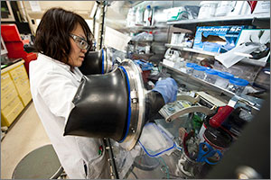 This photo shows a scientist in a white lab coat and with her right arm in a glove box. The glove box has a busy collection of plastic containers.
