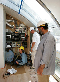 Photo of four men in hard hats. Two of the men are connecting up wires, and the other two are observing, inside a control room of one of the houses that competed in the 2005 Solar Decathlon. 