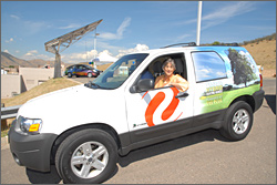 Photo of a smiling woman driving a white SUV vehicle in a parking lot. The side panel reads: Plug-in hybrid electric vehicle, powered at the plug. The Xcel Energy logo is on the door panel.