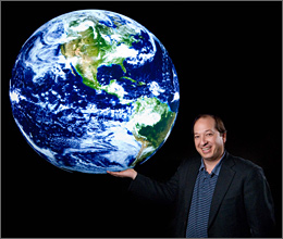 Photo of a man in a sports coat with outstretched arm and palm of hand facing up to make it appear he is holding the Earth in his hand.