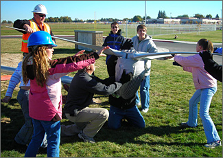 Photo of two adults and five kids outside who are working together to spin a fan-shaped rotor onto a pole.