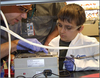 Photo close-up of a man with a goatee beard and a boy wearing a white lab coat; the man points with a gloved hand to a piece of lab experiment.