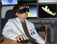 Photo of a boy sitting at a table clicking a computer mouse while wearing a white lab coat and large opaque black 3-D glasses. Behind him on an electronic display wall are brightly colored animated images of molecules.