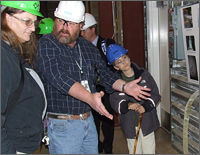 Photo of a bearded man in a plaid shirt and wearing a hard hat speaking with another man with long hair and a boy.  They are looking at photographs of wind turbine experiments mounted on the wall.
