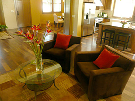 Photo of an interior view of a new bungalow home shows an open floor plan with the parlor in the foreground decorated by two brown chairs and a table with flowers. The kitchen and dining area in the background. 