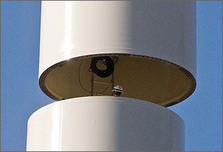 Photo of the head and shoulders of a worker who will connect two large white cylinder-shaped sections of a steel tower. The tower supports a wind turbine.