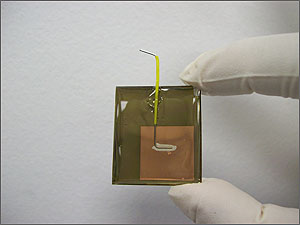 Photo of gloved fingers holding a coin-sized brown square wafer with a wire attached and coming out the top.