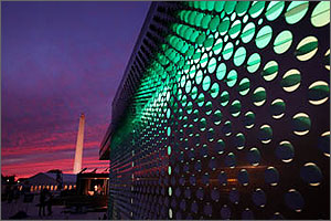 Photo of the contemporary metal exterior of a solar powered house reflecting red and purple light at sunset, with the Washington Monument in the background.