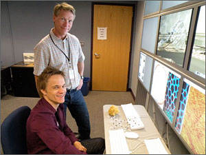Photo of a two men looking at the camera, standing in front of many computer screens.