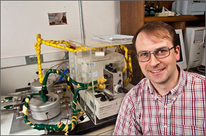 Photo of a man wearing a plaid shirt sitting in front of scientific instruments on a table.